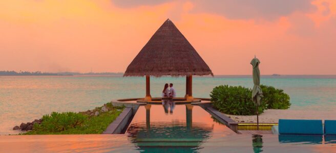 A couple enjoying a romantic moment by the sea and pool under a beach cabana during sunset.